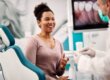 Woman sitting in a dental chair smiling and talking with a dentist wearing a mask, with a screen showing dental images in the background.