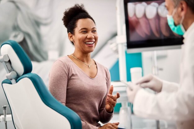 Happy black woman talks to her dentist during appointment at dental clinic. Woman sitting in a dental chair smiling and talking with a dentist wearing a mask, with a screen showing dental images in the background.