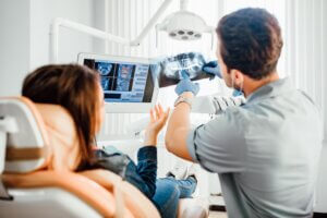 A dentist wearing gloves shows a patient her dental X-rays on film and on a digital monitor during a consultation in a bright, updated dental office.