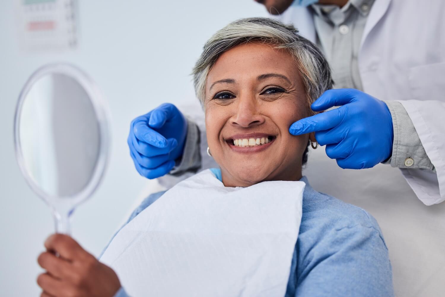 Smiling woman holding a hand mirror while a dentist in blue gloves gently examines her cheeks during a dental appointment.